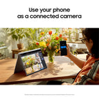 A woman sits at a wooden table in a sunlit indoor setting, holding a smartphone that displays a camera app, while a tablet on a stand shows a video call interface. Large text at the top reads, 'Use your phone as a connected camera.