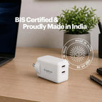 A white Stuffool dual USB-C power adapter sits on a light wood desk next to a laptop, with a potted plant in the background and text overlays reading 'BIS Certified & Proudly Made in India' and a circular 'Made in India' badge.