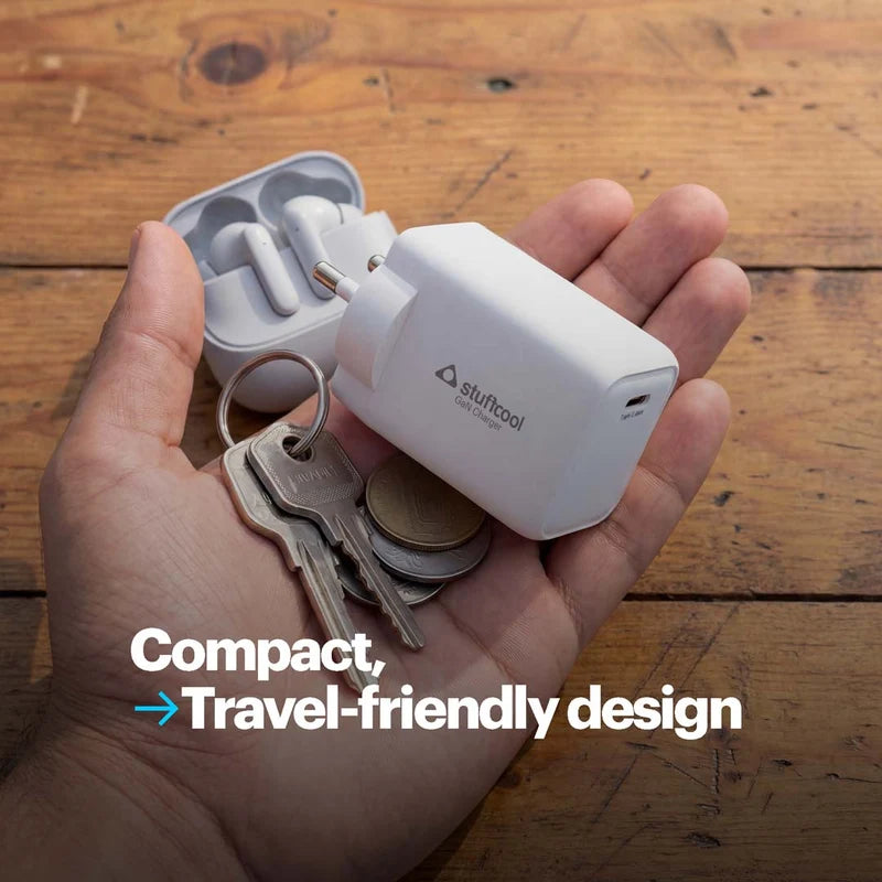 A hand holds a compact white travel charger with a plug and a small white earbud charging case beside it, with keys and coins in the palm. The charger has the stuftool logo printed on it, and the background is a wooden tabletop.