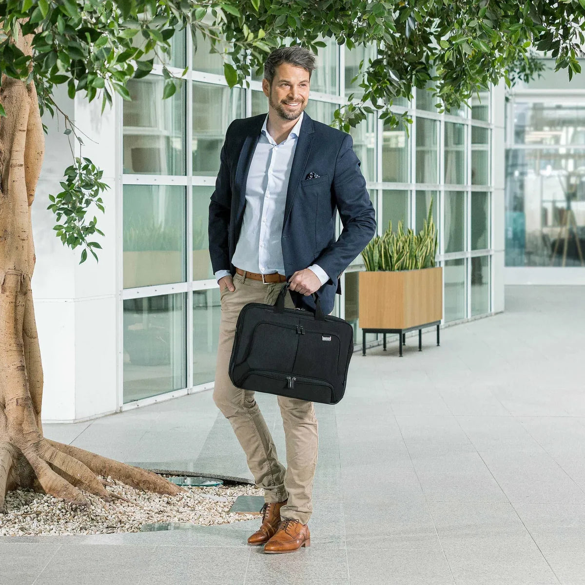Man holding a black briefcase in an indoor setting with large windows and plants.