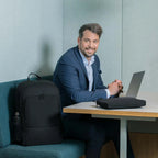 A man in a blue suit sits at a desk with a laptop and a black laptop sleeve, with a matching black backpack resting on the teal bench beside him in an office setting.