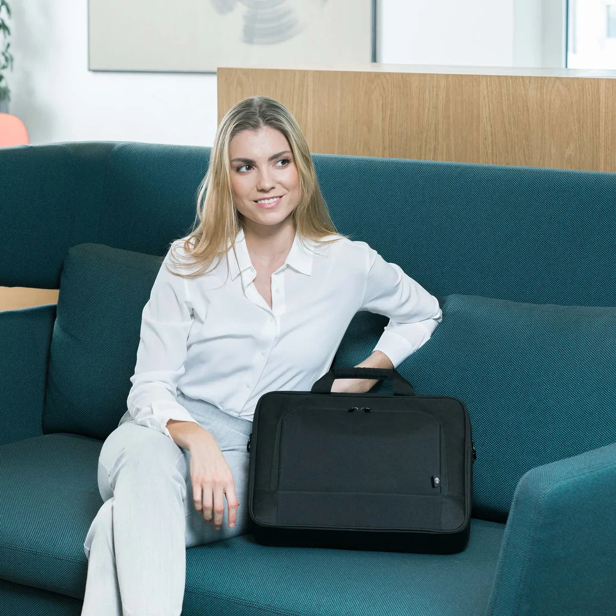 Woman sitting on a teal couch holding a black bag in an indoor setting.