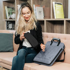 A woman sits on a pink sofa holding a black laptop sleeve, with a gray laptop sleeve resting on the sofa beside her. The setting is a cozy room with wooden shelves in the background.