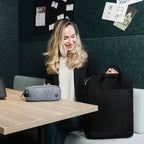 A woman sits at a wooden table in an office setting, holding a black laptop bag next to a gray zippered pouch on the table. The background features a dark green felt wall with pinned papers.