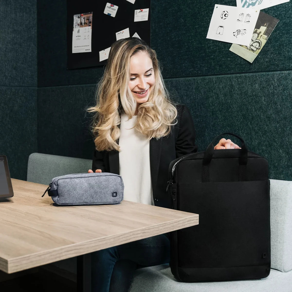 A woman sits at a wooden table in an office setting, holding a black laptop bag next to a gray zippered pouch on the table. The background features a dark green felt wall with pinned papers.