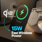 A smartphone shown at an angle on a wireless charging pad in a real indoor setting, with overlaid text reading 'Qi2 Compatible', '15W Fast Wireless Power', and a green battery icon on the phone screen. The product appears to be a wireless charging dock/stand rather than the phone itself.