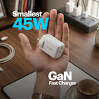 A hand holds a small white GaN fastest charger in its palm, with text overlays reading 'Smallest 45W' and 'GaN Fast Charger'. The background shows a wooden desk with a power strip, cables, and a coffee cup.