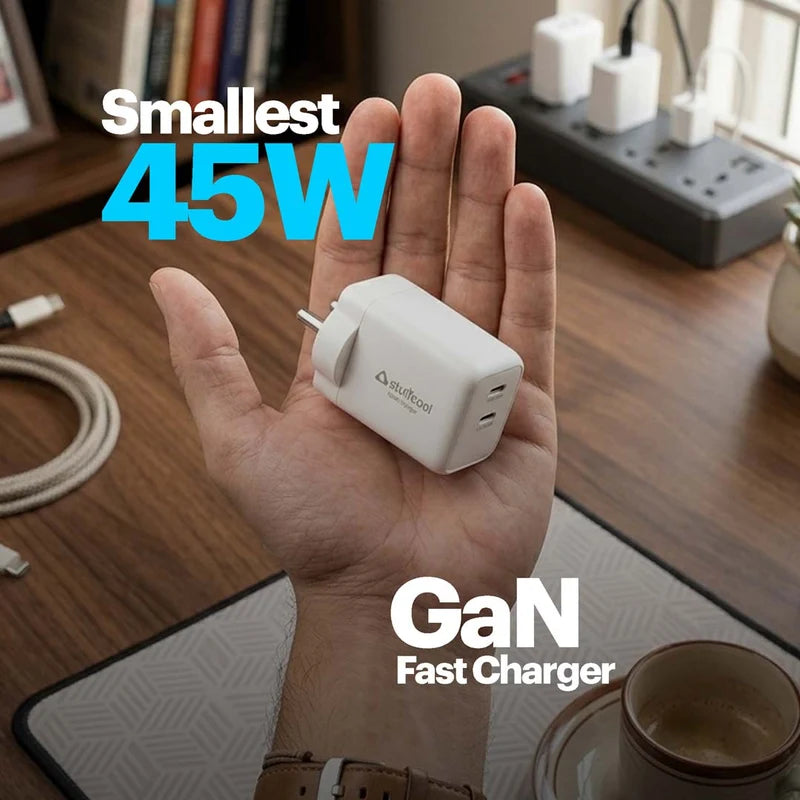 A hand holds a small white GaN fastest charger in its palm, with text overlays reading 'Smallest 45W' and 'GaN Fast Charger'. The background shows a wooden desk with a power strip, cables, and a coffee cup.